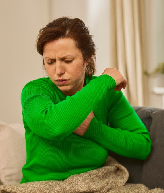 Eine Frau in einem grünen Pullover sitzt auf einem Sofa und hustet in ihren Ellenbogen.