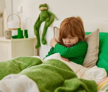 Junges Mädchen mit roten Haaren in einem grünen Oberteil niest in die Armbeuge während es im Bett liegt.