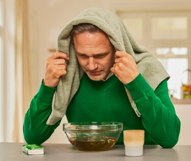 Mann in grünem Pullover mit einem Handtuch über dem Kopf beugt sich über eine Schüssel mit dampfendem Wasser.