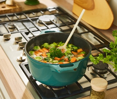 Frische Hühnersuppe köchelt auf einem Herd in einem blauen Topf, umgeben von Zutaten und Küchenutensilien.