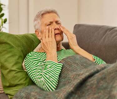 Eine ältere Frau mit grauen Haaren, gekleidet in einem gestreiften grün-weißen Pullover, sitzt auf einem Sofa, hält sich die Handflächen an die Nebenhöhlen und scheint Schmerzen zu empfinden, möglicherweise aufgrund einer Kieferhöhlenentzündung.