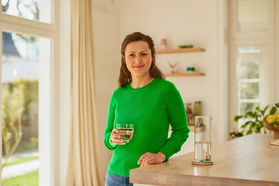 Lächelnde Frau im grünen Pullover steht in einer hellen Küche und hält ein Glas Wasser in der Hand.