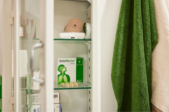 Gelomyrtol Forte – Ein fester Bestandteil der Hausapotheke für die Erkältungs- und Grippesaison. Blick in einen offenen Medizinschrank mit einer Packung Gelomyrtol Forte sichtbar auf einem Regal, neben anderen Gesundheitsprodukten und einem grünen Handtuch an der Tür hängend.