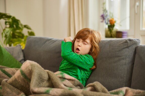 Kind hustet auf dem Sofa – Darstellung von Erkältung und spastischer Bronchitis bei Kindern. Kleines Mädchen mit rotem Haar hustet auf einem Sofa, eingewickelt in eine Decke, in einem wohnlichen Raum. Darstellung von Husten und Erkältungssymptomen bei Kindern.