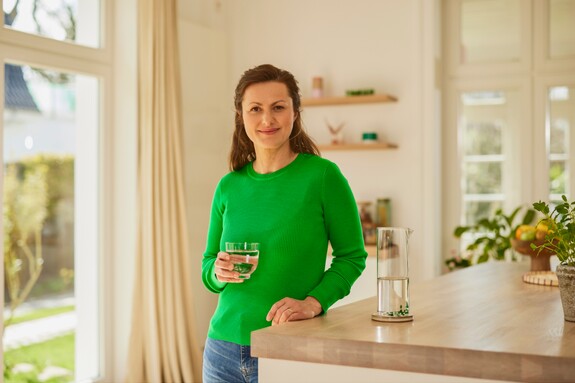 Gesunde Frau in der Küche mit einem Glas Wasser - Gelomyrtol Forte unterstützt Ihre Gesundheit. Lächelnde Frau im grünen Pullover steht in einer hellen Küche und hält ein Glas Wasser in der Hand.