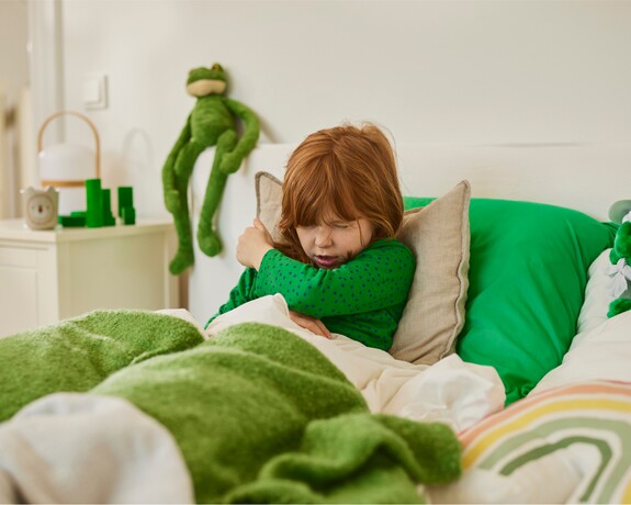 Gelomyrtol forte – Sanfte Linderung für Kinder bei Bronchitis und Erkältungssymptomen. Junges Mädchen mit roten Haaren in einem grünen Oberteil niest in die Armbeuge während es im Bett liegt.