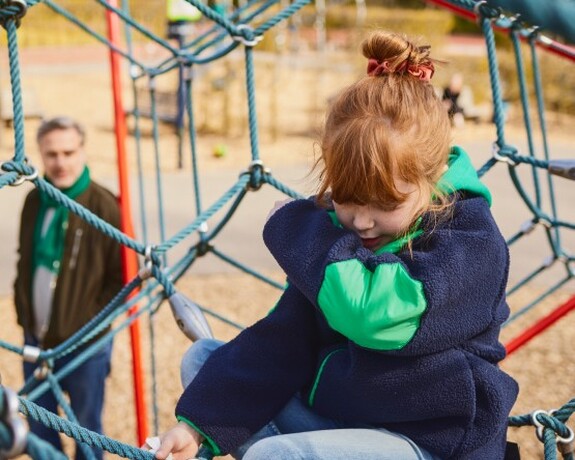 Gelomyrtol Forte – Unterstützung der Gesundheit von Kindern, damit sie aktiv bleiben und das Spielen im Freien genießen können. Rothaariges Mädchen sitzt auf einem Klettergerüst und hustet sich in die Armbeuge während der Vater im Hintergrund steht und sie beobachtet.