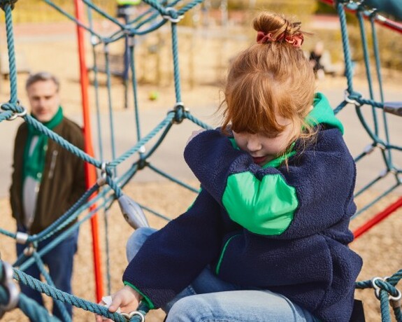 Gelomyrtol Forte – Unterstützung der Gesundheit von Kindern, damit sie aktiv bleiben und das Spielen im Freien genießen können. Rothaariges Mädchen sitzt auf einem Klettergerüst und hustet sich in die Armbeuge während der Vater im Hintergrund steht und sie beobachtet.