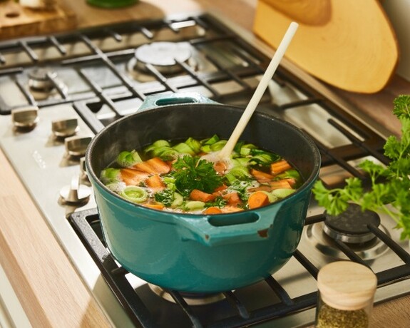 Gelomyrtol Forte – Ergänzung zu einer ausgewogenen Ernährung für eine bessere Gesundheit und Erkältungsvorbeugung. Frische Hühnersuppe köchelt auf einem Herd in einem blauen Topf, umgeben von Zutaten und Küchenutensilien.